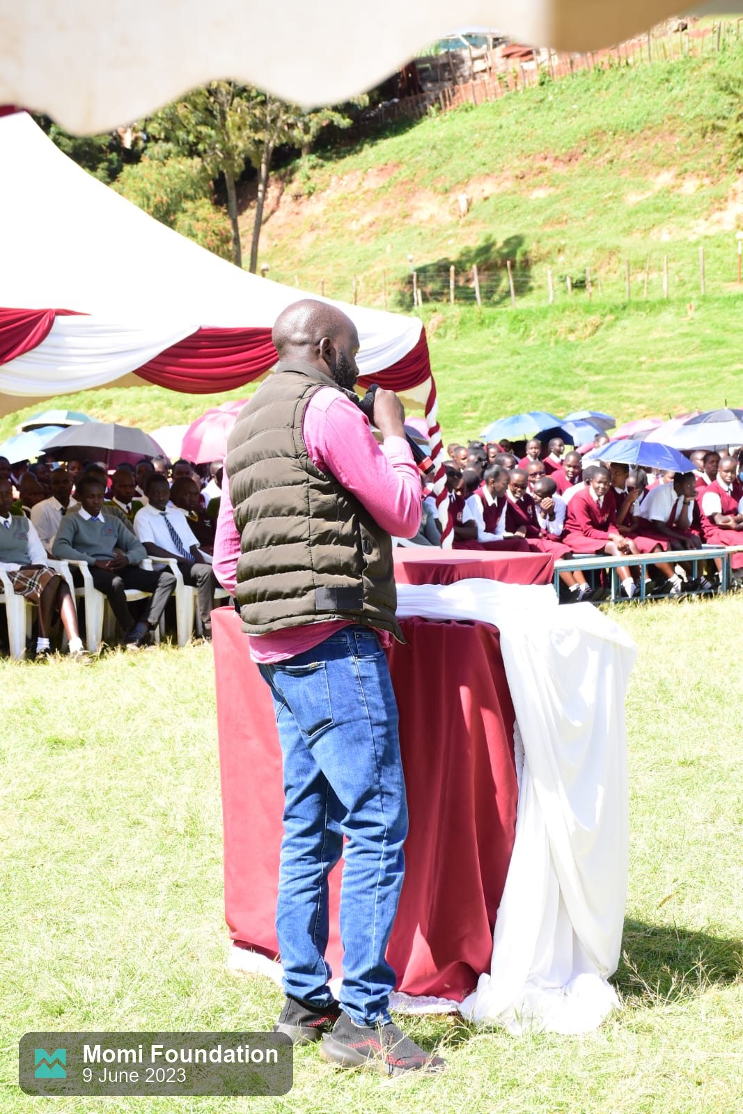 A speaker addresses rows of students seated under umbrellas